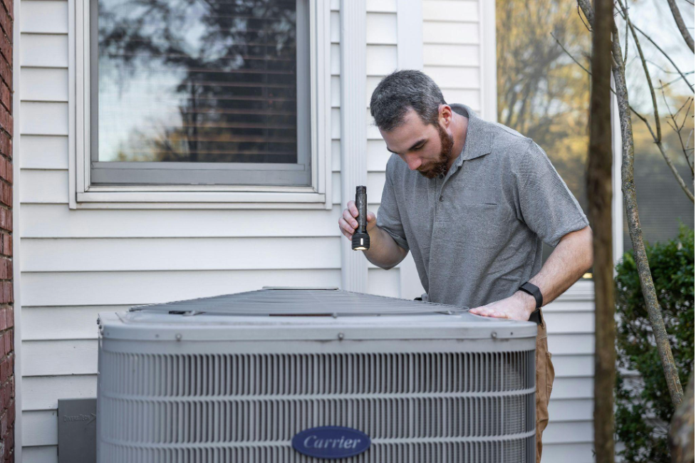 A person shining a flashlight inside a central air conditioning unit outside a home.