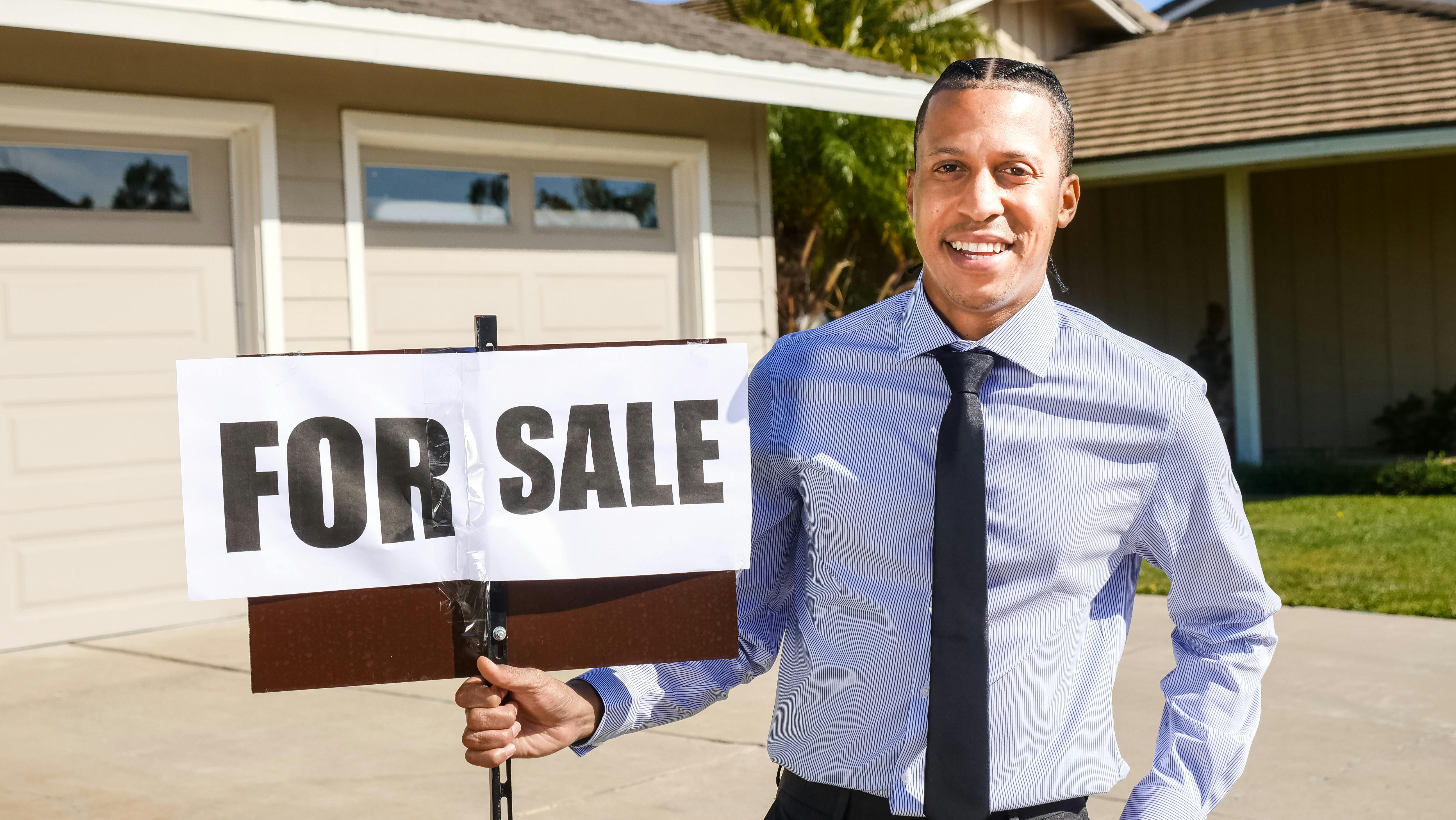a man holding a “for sale” sign in front of a garage